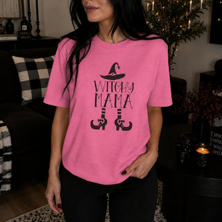 A woman wearing a pink short-sleeve t-shirt with a black graphic that reads 'WITCHY MAMA' with a witch hat and boots, in a cozy, dimly lit living room setting.