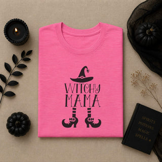 A folded bright pink T-shirt with a black witch-themed graphic reading 'WITCHY MAMA' and showing a witch hat and high heels, styled on a beige fabric background with black candles, pumpkins, dried stems, and a black book.