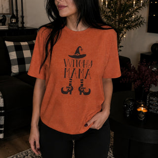 A woman wearing a heather orange short-sleeve t-shirt with a black graphic that reads 'WITCHY MAMA' with a witch hat and witch boots, standing in a cozy, dimly lit living room.
