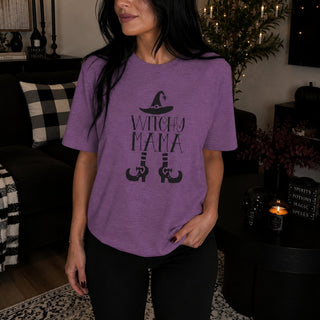 A woman wearing a heather purple graphic t-shirt with a black 'WITCHY MAMA' and witch hat design, standing in a cozy, dimly lit living room.