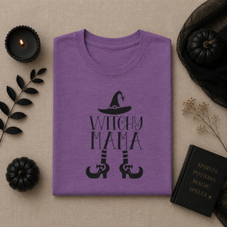 A folded purple t-shirt with a black 'Witchy Mama' graphic featuring a witch hat, text, and boot prints, styled in a flat lay on a beige linen background with black pumpkins, a candle, dried flowers, and a black book.