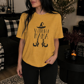 A woman wearing a heather mustard yellow graphic t-shirt with a black 'WITCHY MAMA' text design and witch hat/boots illustration, paired with black pants in a cozy, dimly lit living room setting.