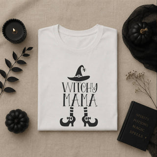 A folded white T-shirt with a black 'WITCHY MAMA' graphic and witch hat/stocking design, styled in a flat lay on a beige fabric background with black candles, pumpkins, dried stems, and a black book.