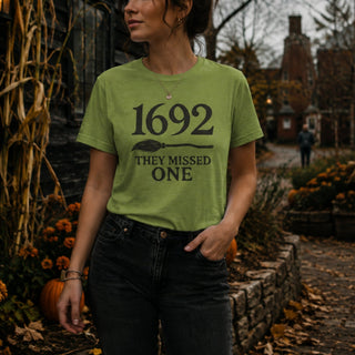 A woman wearing a sage green graphic t-shirt with the text '1692 THEY MISSED ONE' printed in black, standing outdoors in a rustic autumn setting with pumpkins and a stone path.