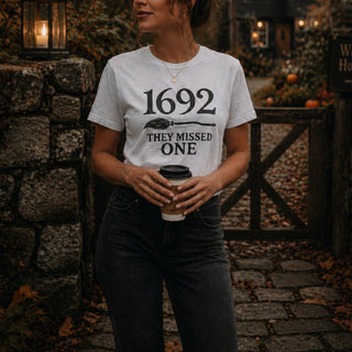 A woman wearing a white graphic t-shirt with the text '1692 THEY MISSED ONE' printed in black, standing in an outdoor autumn setting and holding a coffee cup.