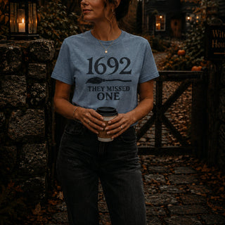 A woman wearing a heathered blue graphic t-shirt with the text '1692 THEY MISSED ONE' and a spoon design, holding a coffee cup in a rustic outdoor setting.