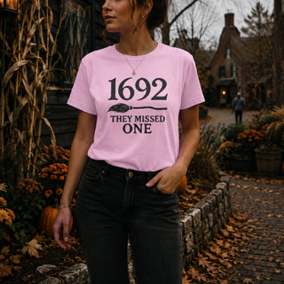 A woman wearing a light pink graphic t-shirt with the black text '1692 THEY MISSED ONE' and a witch's broom graphic, paired with black jeans, standing in an outdoor autumn yard.