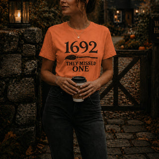 A woman wearing an orange graphic t-shirt with the text '1692 THEY MISSED ONE' printed in black, holding a coffee cup in a rustic outdoor setting.
