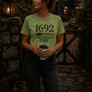 A woman wearing a sage green graphic t-shirt with the text '1692 THEY MISSED ONE' and a spoon graphic, standing in a rustic outdoor setting near a stone wall and wooden gate.