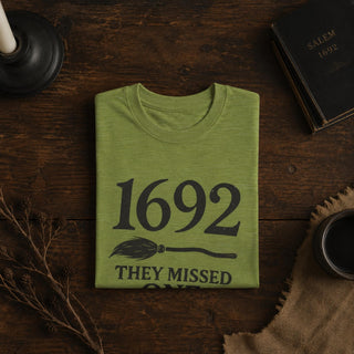 A folded sage green t-shirt with a black graphic print reading '1692 THEY MISSED ONE' and a broomstick illustration, displayed on a dark wooden tabletop with styled flat-lay props including a candle, book, and mug.