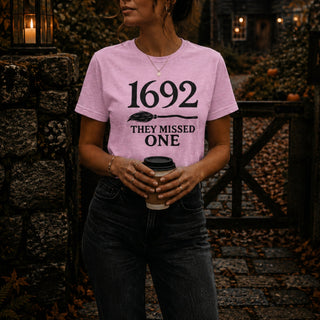 A woman wearing a light pink graphic t-shirt with the text '1692 THEY MISSED ONE' and a broomstick design, holding a coffee cup in an outdoor autumn setting.