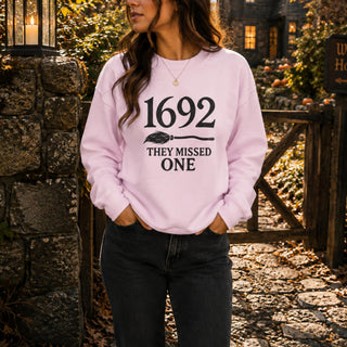A woman wearing a light pink crewneck sweatshirt with the black printed text '1692 THEY MISSED ONE' and a broom graphic, standing outdoors in front of a stone wall and wooden gate.