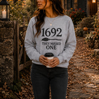 A woman wearing a light gray crewneck sweatshirt with a black graphic that reads '1692 THEY MISSED ONE' and holding a coffee cup, standing on a leaf-covered outdoor path.