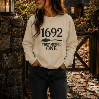 A woman wearing a cream-colored crewneck sweatshirt with a black graphic that reads '1692 THEY MISSED ONE' under a broom icon, standing outdoors in front of a rustic stone wall and wooden gate.