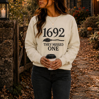 A woman wearing a cream-colored crewneck sweatshirt with a black graphic print that reads '1692 THEY MISSED ONE' with a broomstick illustration. She is holding a white coffee cup and standing outdoors on a stone path surrounded by autumn leaves.
