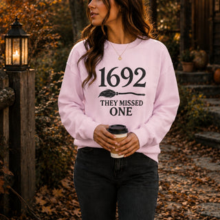 A woman wearing a light pink crewneck sweatshirt with a black graphic print that reads '1692 THEY MISSED ONE' and holding a coffee cup in an outdoor autumn setting.