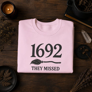 A folded light pink crewneck t-shirt with a black graphic reading '1692 THEY MISSED' and a broom illustration, displayed on a dark wooden surface with candles, dried botanicals, and crystals around it.