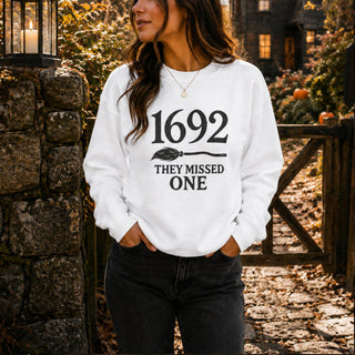 A woman wearing a white crewneck sweatshirt with a black graphic that reads '1692 THEY MISSED ONE' with a broomstick illustration, standing outdoors in front of a stone wall and wooden gate.