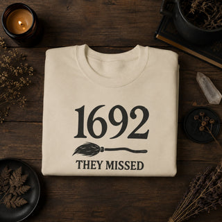 A folded cream-colored t-shirt with a black graphic reading '1692 THEY MISSED' and a broomstick illustration, displayed on a dark wooden table surrounded by dried botanicals, a candle, and books.