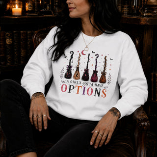 A woman wearing a white crewneck sweatshirt with a graphic of six guitars and the text 'A GIRL'S GOTTA HAVE OPTIONS' in colorful lettering, seated in a dark wooden armchair.