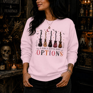 A woman wearing a light pink crewneck sweatshirt with a graphic of six guitars and the text 'A GIRL'S GOTTA HAVE OPTIONS' in a dark, gothic-themed shop setting.
