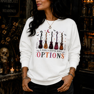 A woman wearing a white crewneck sweatshirt with a graphic of six guitars and the text 'A GIRL'S GOTTA HAVE ALL OPTIONS' in a dark, atmospheric shop setting.