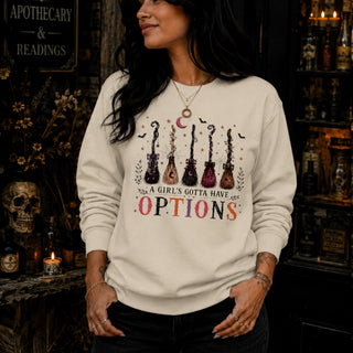 A woman wearing a cream-colored sweatshirt with a graphic of five guitars and the text 'A GIRL'S GOTTA HAVE OPTIONS' in pink and black, posed in a dark, atmospheric shop setting.