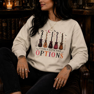A woman wearing a cream-colored crewneck sweatshirt with a graphic of six guitars and the text 'A GIRL'S GOTTA HAVE OPTIONS' in colorful lettering. She is sitting in a dark wooden chair in a cozy, dimly lit room with bookshelves in the background.