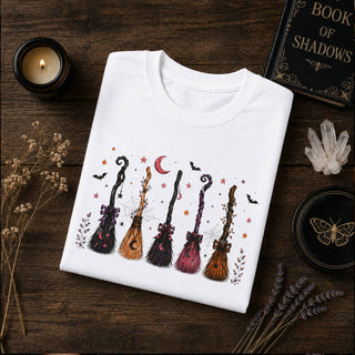 A folded white crewneck sweatshirt with a colorful Halloween-themed broomstick graphic, styled on a dark wooden table with a candle, a book, crystals, and dried flowers.