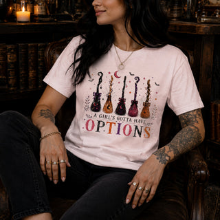 A woman wearing a light pink graphic t-shirt with a design of five guitars and the text 'A GIRL'S GOTTA HAVE OPTIONS', seated in a dark leather armchair in a dimly lit room with bookshelves in the background.