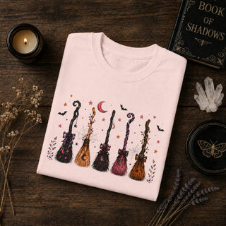 A folded light pink crewneck sweatshirt with a Halloween-themed graphic print of five broomsticks, stars, bats, and a crescent moon, displayed on a dark wooden background with decorative props including a candle, book, and dried flowers.