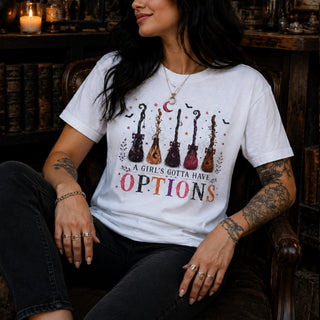 A woman wearing a white graphic t-shirt with a colorful illustration of six guitars and the text 'A GIRL'S GOTTA HAVE OPTIONS', seated in a dark wooden chair in a cozy, dimly lit room.