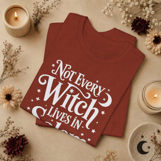 A folded terracotta red t-shirt with a white graphic reading 'Not Every Witch Lives In Ohio' surrounded by stars and crescent moons, styled in a cozy flat-lay with candles, dried flowers, and a ceramic dish on a beige background.
