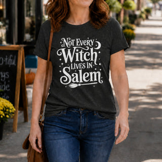 A woman wearing a dark charcoal gray graphic t-shirt with the white text 'Not Every Witch Lives in Salem' and a broomstick design, paired with blue jeans, walking on a sunny sidewalk.