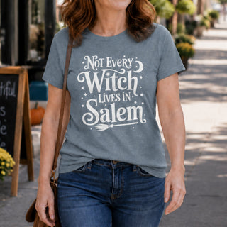 A woman wearing a heather slate blue graphic t-shirt with the white text 'Not Every Witch Lives in Salem' and a broomstick design, paired with blue jeans, walking on a sunny outdoor street.