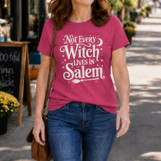 A woman wearing a pink graphic t-shirt with the white text 'Not Every Witch Lives in Salem' and blue jeans, walking on a sunny outdoor street.