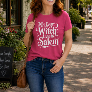 A woman wearing a bright pink graphic t-shirt with the white text 'Not Every Witch Lives in Salem' and blue jeans, carrying a brown leather shoulder bag in an outdoor shopfront setting.
