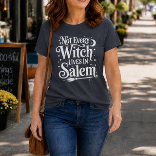 A woman walking on a sunny outdoor sidewalk wearing a dark gray graphic t-shirt with the white text 'Not Every Witch Lives in Salem' and a broomstick design, paired with blue jeans.