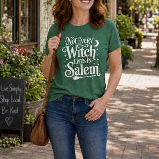A woman wearing a green graphic t-shirt with the text 'Not Every Witch Lives in Salem' in white, paired with blue jeans and carrying a brown shoulder bag, standing on a sunny brick sidewalk.