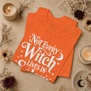 An orange folded graphic T-shirt with white text reading 'Not Every Witch Lives In' surrounded by decorative dried flowers, candles, and a ceramic dish on a neutral beige surface.