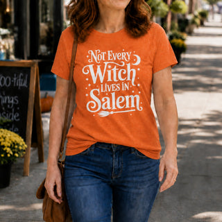 A woman wearing an orange graphic t-shirt with the white text 'Not Every Witch Lives in Salem' and a broomstick design, paired with blue jeans, walking on a sunny sidewalk.