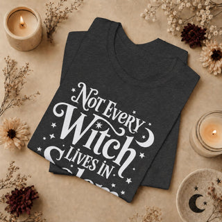 A dark charcoal gray t-shirt with a white graphic print reading 'Not Every Witch Lives In Fear' is displayed in a flat-lay arrangement on a beige background, surrounded by dried flowers, candles, and a ceramic plate.