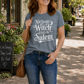 A woman wearing a heather gray graphic t-shirt with the white text 'Not Every Witch Lives in Salem' and blue jeans, standing on a sunny brick sidewalk in a shop street.