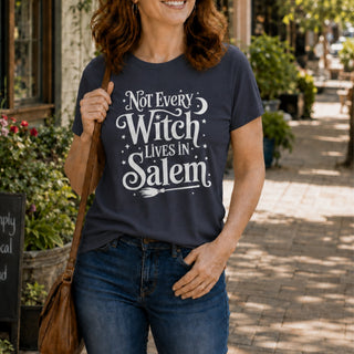 A woman wearing a navy blue graphic t-shirt with the white text 'Not Every Witch Lives in Salem' and a broomstick design, paired with blue jeans, standing on a sunlit brick sidewalk.
