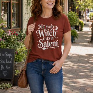 A woman wearing a rust-brown graphic t-shirt with the white text 'Not Every Witch Lives in Salem' and blue jeans, standing on a sunny outdoor brick sidewalk.