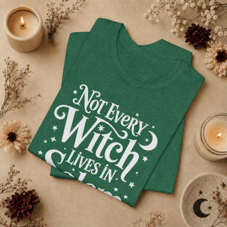 A folded green graphic T-shirt with white text reading 'Not Every Witch Lives In' is styled in a flat lay on a beige surface with dried flowers, candles, and a small ceramic dish.