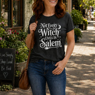 A woman wearing a dark charcoal gray graphic t-shirt with the white text 'Not Every Witch Lives In Salem' and a broomstick illustration, paired with blue jeans and carrying a brown shoulder bag in an outdoor street setting.