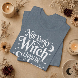 A folded heather slate blue t-shirt with a white graphic reading 'Not Every Witch Lives In Peace' surrounded by stars and crescent moons, styled in a flat lay with dried flowers, candles, and a ceramic dish on a beige background.