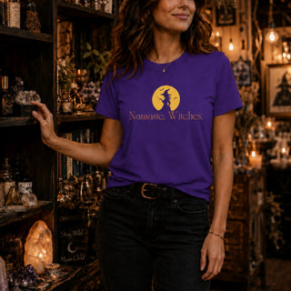 A woman wearing a purple graphic t-shirt with a gold 'Namaste, Witches' design, standing in a cozy, dimly lit shop filled with crystals and books.