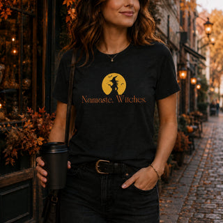 A woman wearing a black graphic t-shirt with a yellow witch silhouette and the text 'Namaste, Witches' on the front, standing on a cobblestone street.